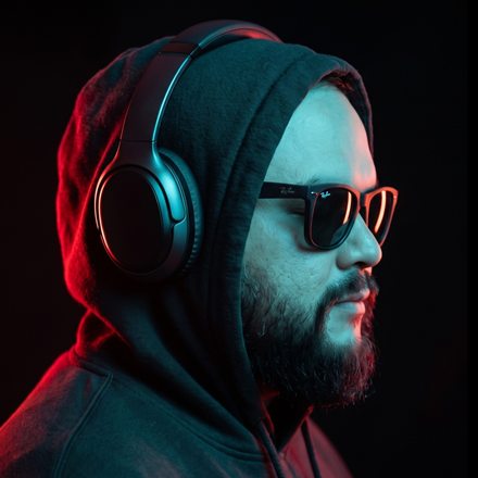 Jonathan con audífonos, capucha y lentes oscuros, iluminado en rojo y turquesa