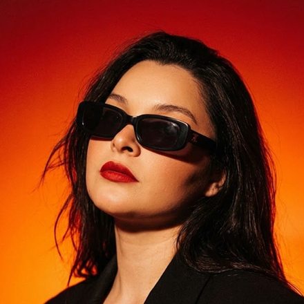 Maye con lentes oscuros y labios rojos sobre fondo naranja